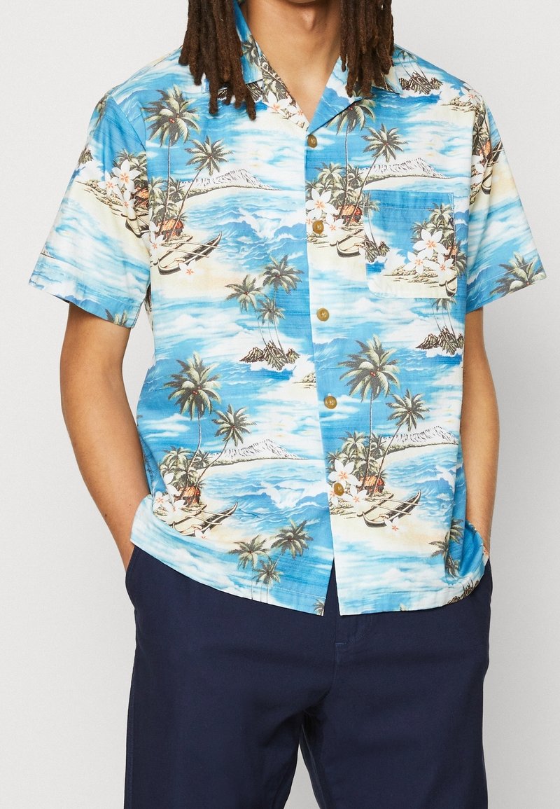 Homme portant une chemise hawaïenne bleue à manches courtes avec un motif de palmiers, bateaux et océan, associée à un pantalon bleu foncé, mains dans les poches.