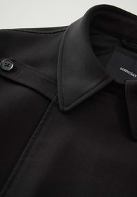 Zwarte jacket met een kraagontwerp, knoopsluiting en een gladde stoftextuur. Prominente stiksels langs de naden.