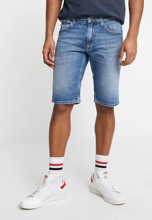 Shorts en denim bleu clair avec une coupe décontractée, comportant cinq poches, un bouton métallique et des ourlets cousus. Associés à des baskets blanches et des chaussettes rayées.