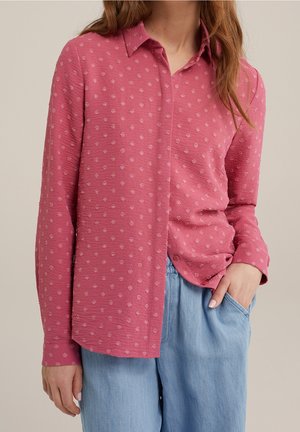 Kvinde iført en langærmet pink tekstureret skjorte med små prikker og lyseblå løse bukser, står med den ene hånd i lommen.