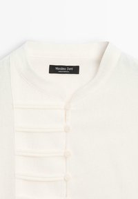 Weißes, strukturiertes Kleidungsstück mit rundem Ausschnitt, das eine Knopfleiste und vertikale Falten hat. Das Etikett trägt die Aufschrift "Massimo Dutti, Made in Portugal."