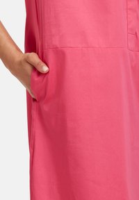 Rosa Stoff mit einer glatten Textur und einer Seitentasche, gezeigt mit einer Hand, die in der Tasche ruht, wodurch das Design des Kleidungsstücks hervorgehoben wird.