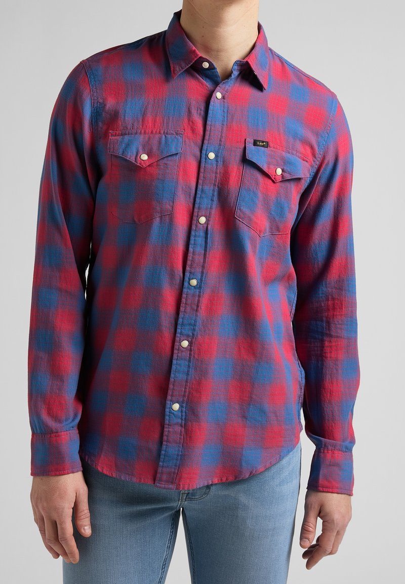 Homme portant une chemise à carreaux rouge et bleue à manches longues avec des poches poitrine boutonnées et un jean bleu clair.