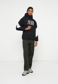 Juoda ir balta gobtuvinė striukė su užrašu "NIKE AIR" priekyje, derinama su alyvuogių žalumo cargo kelnėmis ir pilkais sportbačiais.