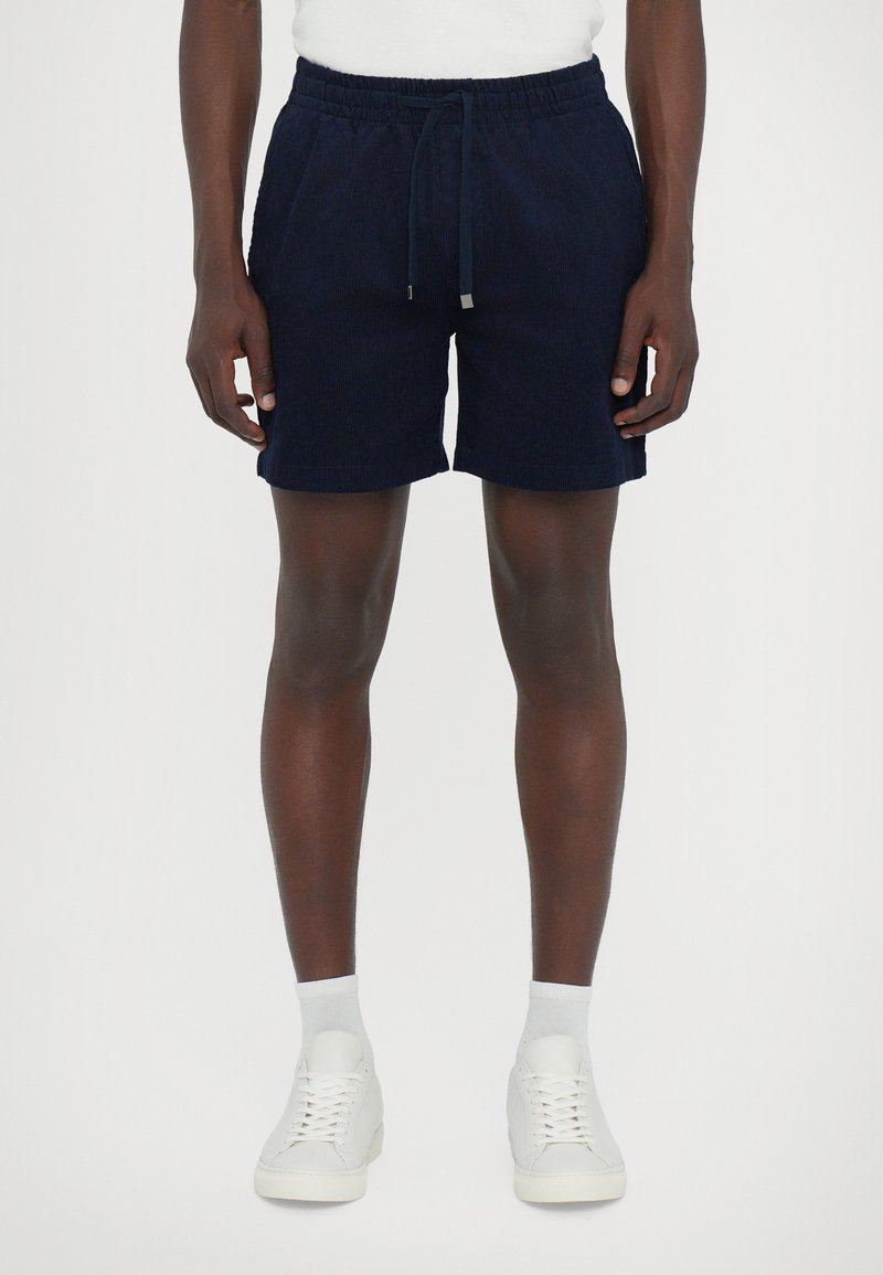 Shorts en velours côtelé bleu marine avec une taille élastique, un cordon de serrage et deux poches latérales. Texture lisse et coupe décontractée. Baskets et chaussettes blanches.