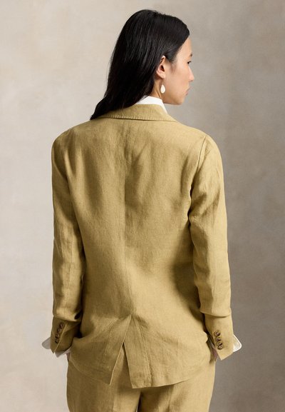 Mustade juustega naine, kes kannab heledat pruuni linaülikonda, vaadatuna tagantpoolt, neutraalse beeži tausta taustal.