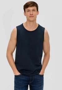 Young man standing with hands in pockets, wearing a navy sleeveless shirt and dark blue jeans against a plain light background.