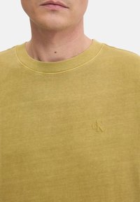Camiseta de algodón color mostaza con cuello redondo. Presenta un sutil logo "CK" en relieve en la parte delantera. Textura suave y corte clásico.