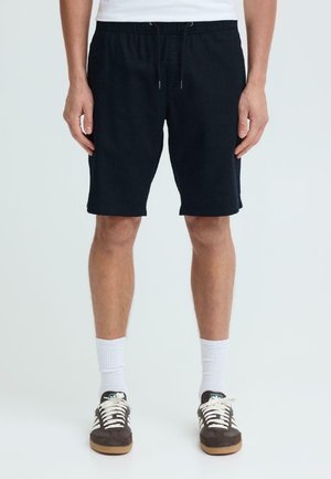 Mann trägt schwarze Shorts mit Kordelzug, weiße Socken und braune Sneakers, steht vor weißem Hintergrund.