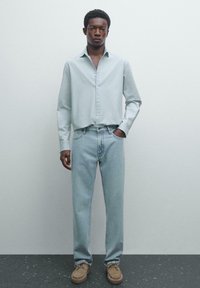 Uomo in piedi contro un muro semplice, indossa una camicia a maniche lunghe azzurro chiaro, jeans azzurri chiari e scarpe in suede beige, con una mano in tasca.