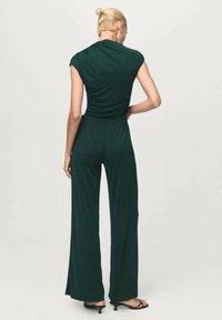 Temno zelena kombinezona s kratkimi rokavi, prilagojenim telesom in širokimi hlačnicami. Gladek, elastičen material z minimalnimi detajli.