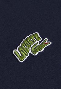 Marinblått tyg med en grön broderad "LACOSTE"-logotyp med en krokodilaccent, inramad i vitt, vilket visar textur och varumärkesdetaljer.