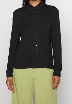 Chemise noire à manches longues avec un col, fermeture à boutons sur le devant et texture douce. Associée à un pantalon vert clair, mettant en avant une coupe ample.