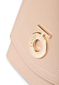 Beige nahkalaukku kultaisilla yksityiskohdilla, jossa on pyöreä logo ja kontrastivärinen valkoiset tikkaus yksityiskohtaisessa rakenteessa.