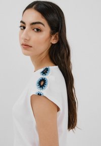 Hvid t-shirt med korte ærmer, der har blå og sorte broderede blomsterdetaljer på skuldrene. Glat tekstur og enkelt design.