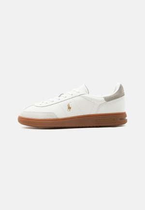 Biały niski sneaker z brązową podeszwą, zamszowymi akcentami na palcach i pięcie, złote logo Polo po boku, sznurowany, gumowa podeszwa z oznaczeniem Ralph Lauren.