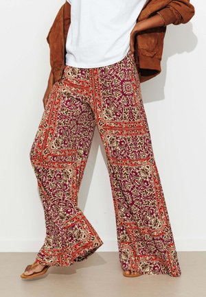 Personne portant un pantalon large avec des motifs complexes rouges, violets et crème, une chemise blanche, une veste marron drapée sur les épaules et des sandales.