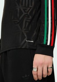 Main portant deux bagues en argent à côté d'une tenue de sport noire ClimaCool avec des bandes vertes, blanches et rouges sur la manche.