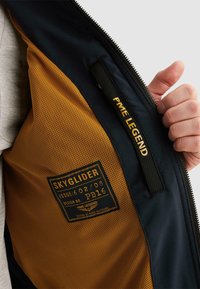 Ruka držící rozepnutou bundu, která odhaluje oranžovou síťovanou podšívku s černou značkou "SKYGLIDER" a vnitřní kapsu s visačkou "PME LEGEND".