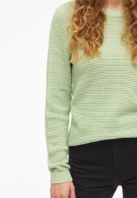 Ανοιχτό πράσινο πλεκτό πουλόβερ με ανάγλυφο σχέδιο, μακριά μανίκια και ριμπ μανσέτες. Συνδυάζεται με σκούρο παντελόνι, εμφανίζοντας άνετη εφαρμογή.