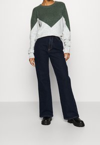 Maglione grigio e verde con un design a zigzag, abbinato a jeans a gamba larga blu scuro e stivali neri chunky. Tessuto testurizzato, vestibilità casual.
