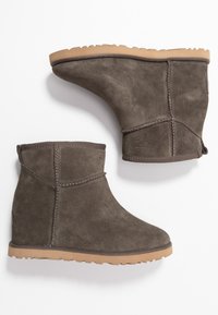 Un par de botas de ante gris a nivel del tobillo con suelas de goma marrón, costuras visibles y una lengüeta en la parte trasera, exhibidas sobre un fondo blanco.