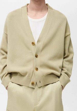 Beżowy kardigan na guziki noszony na białej koszulce, zestawiony z jasnobeżowymi spodniami, ręce wsunięte w kieszenie.