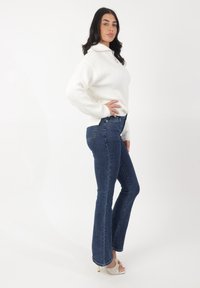 Pull blanc à côtes avec un col zippé mi-haut, associé à un jean évasé bleu foncé. La tenue est rehaussée par des chaussures à talons hauts à lanières.
