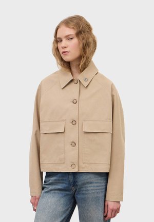 Młoda osoba ubrana w beżową zapinaną na guziki kurtkę z dużymi przednimi kieszeniami i niebieskie jeansy, stojąca na tle jednolitego jasnego tła.