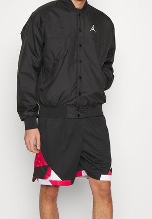 Mann trägt eine schwarze Jacke mit Knöpfen und weißem Logo sowie schwarze Sportshorts mit roten, weißen und schwarzen geometrischen Mustern.