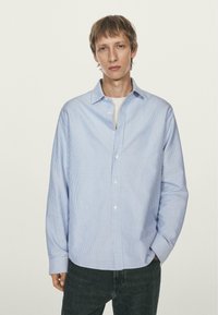 Giovane uomo che indossa una camicia button-up a righe azzurre su una maglietta bianca, con jeans scuri, in piedi davanti a uno sfondo chiaro e semplice.