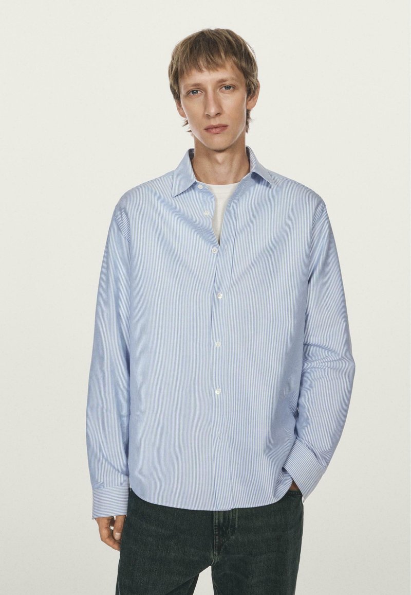 Giovane uomo che indossa una camicia button-up a righe azzurre su una maglietta bianca, con jeans scuri, in piedi davanti a uno sfondo chiaro e semplice.