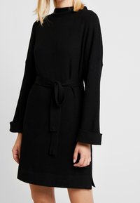 Svart ribbad stickad klänning med långa ärmar, bälte i midjan och sidofickor. Har en hög halsringning och breda manschetter. Mjuk textur.