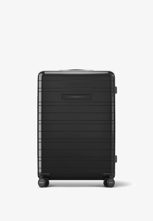 Valise rigide noire avec des rainures horizontales, texture lisse, poignée supérieure et quatre roues pour la mobilité. Le devant présente une poche à glissière.