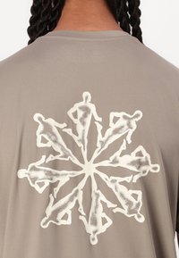 Kortermet, lys taupe T-skjorte med et sentralt hvitt grafikk av menneskefigurer arrangert i et snøflakmønster. Glatt tekstur.