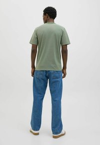 Camiseta de algodón verde de manga corta, ajuste relajado, mostrada con jeans azules y zapatillas blancas, vista desde atrás sobre un fondo claro.