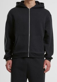 Uomo che indossa una felpa nera con zip e tasche frontali, abbinata a pantaloni neri, su uno sfondo grigio chiaro.