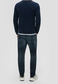 Mies seisoo selin tummansininen villapaita yllä, vaaleat siniset farkut takataskuilla ja vaaleat lenkkarit jalassa.