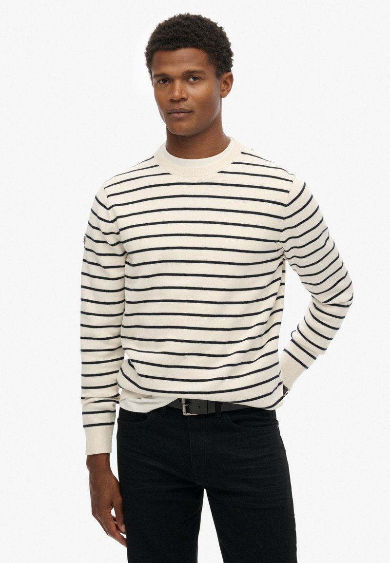 Pullover in Creme mit marineblauen horizontalen Streifen, geripptem Rundhalsausschnitt und Bündchen, figurbetonte Silhouette, kombiniert mit schwarzer Jeans.