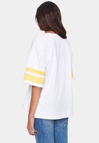 Žena s hnědými vlasy po ramena, oblečená v bílém oversized tričku se žlutými pruhy na rukávech a modrých džínách, otočená zády.