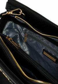 Interieur van een zwarte leren tas met gouden ritsen en hardware. Gevoerd met lichtgrijs stof met een subtiel patroon. Inclusief ritscompartimenten.