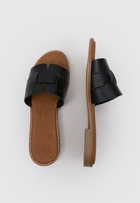 Sandales à glissière en cuir noir avec une large sangle texturée et une semelle intérieure lisse marron. Semelle plate avec un détail de couture de bord de couleur claire.