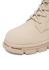 Beżowy skórzany but z fakturowaną powierzchnią, syntetycznymi sznurówkami i złotym metalicznym akcentem na podeszwie, występujący w masywnej, solidnej konstrukcji.