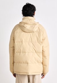 Beige puffer jacket with a hood, featuring horizontal stitched panels, a slightly shiny texture, and elastic cuffs. Back view.