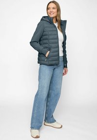 Frau steht und trägt eine dunkelblaue gesteppte Jacke mit Kapuze, ein weißes T-Shirt, helle weitbeinige Jeans und weiße Sneaker, lächelt leicht zur Seite.