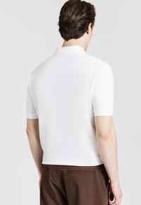 Polo shirt bianca a maniche corte con un motivo testurizzato, caratterizzata da un colletto classico e una silhouette aderente, abbinata a pantaloni marroni.