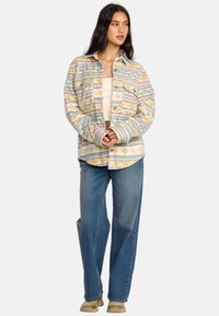 Beige und pastellfarbener Flanellshirt mit horizontalen und welligen Mustern, Knopfleiste vorne, zwei Brusttaschen, getragen über einem weißen Oberteil und blauen Jeans.