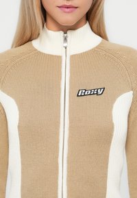Beige strikket hettegenser med høyt krage, med hvite teksturerte sidestykker og et svart logo-patch på brystet.