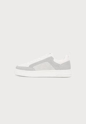 Lavtop hvid sneaker med lysegrå ruskindspaneler, hvide snørebånd og en tyk hvid sål på en ensfarvet lys baggrund.