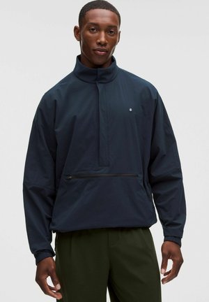 Mann trägt eine marineblaue Pulloverjacke mit einer vorderen Reißverschlusstasche und olivgrüner Hose vor einem schlichten hellen Hintergrund.
